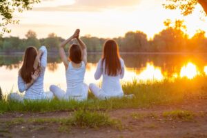 Three women dressed in white engage in Ayurveda Yoga near a lakeside.