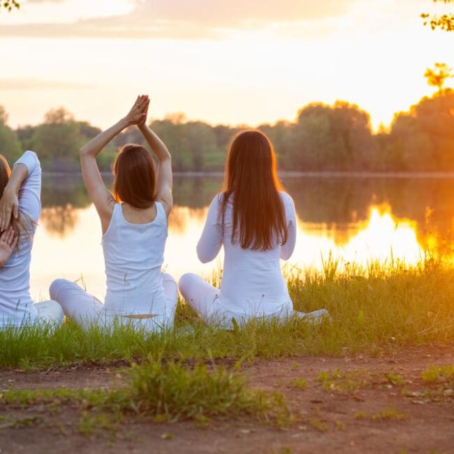 Three women dressed in white engage in Ayurveda Yoga near a lakeside.