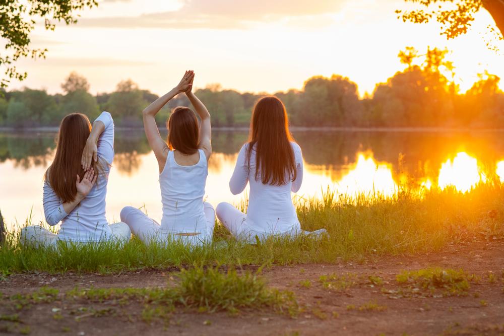 Three women dressed in white engage in Ayurveda Yoga near a lakeside.