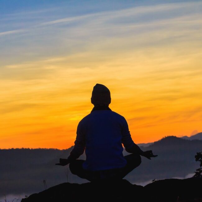 A person meditating on a mountain top with the sunrising around them.