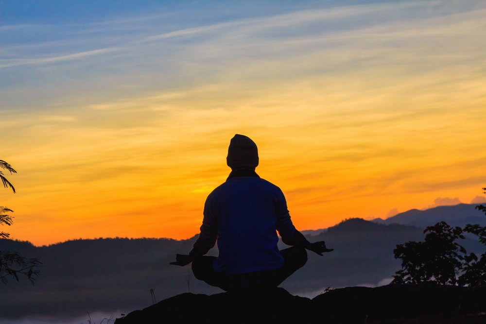A person meditating on a mountain top with the sunrising around them.