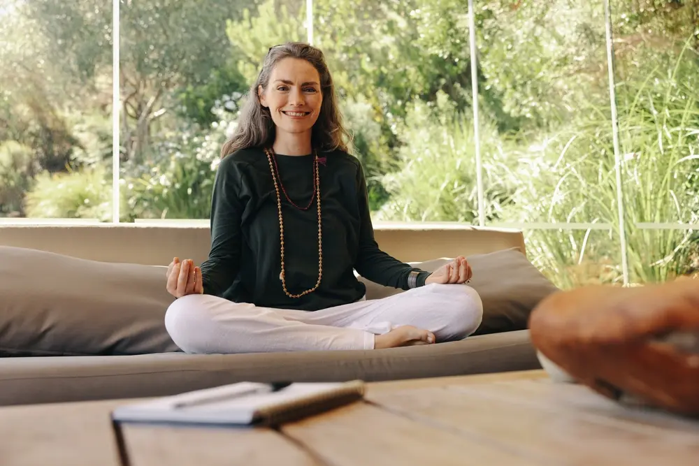 A woman engages in a Vedic meditation practice in front of a window to nature.