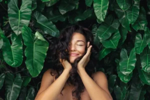 A woman with curly brown hair cupping her face and smiling with her eyes closed in front of greenery.
