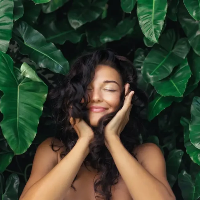 A woman with curly brown hair cupping her face and smiling with her eyes closed in front of greenery.