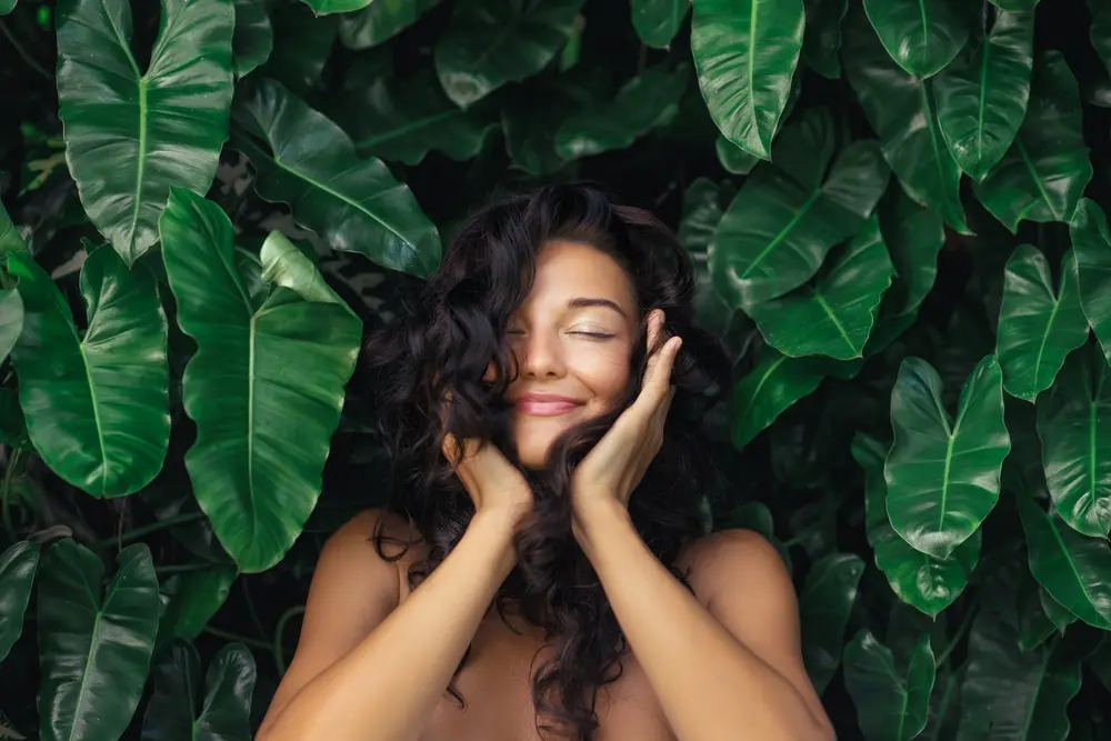 A woman with curly brown hair cupping her face and smiling with her eyes closed in front of greenery.